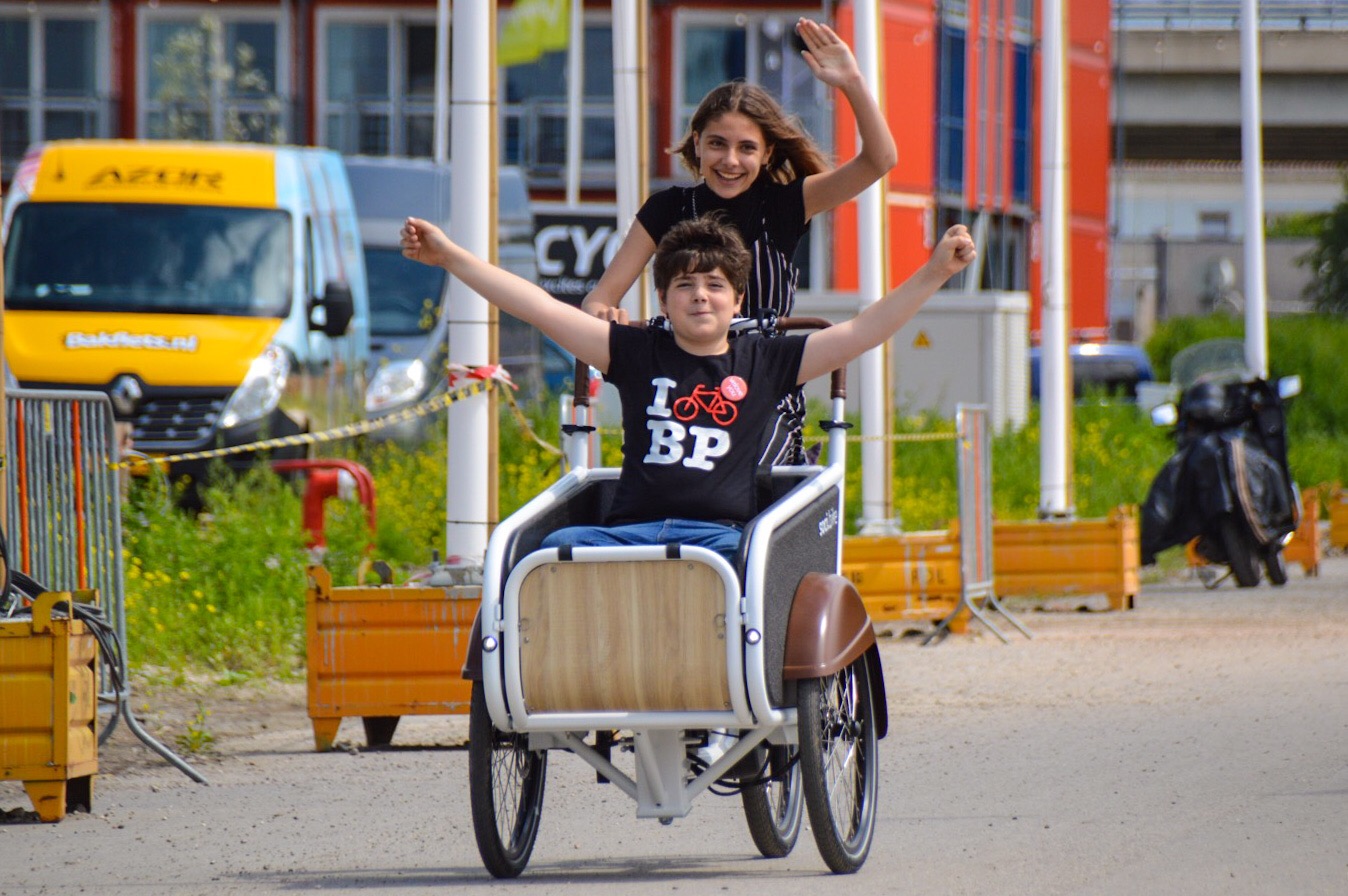 Girl biking a bakfiets while a boy sits in it, both of them smiling!